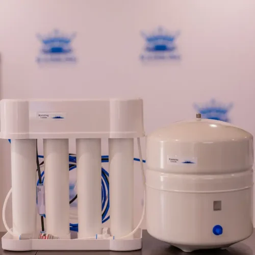 A white water filtration system with multiple cylindrical filters is next to a round white storage tank on a wooden table.